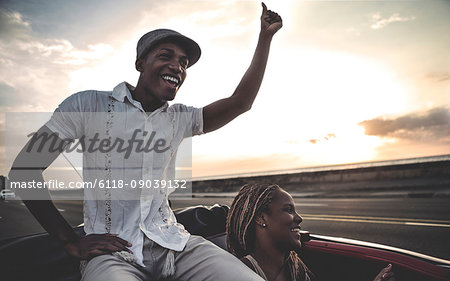 Two people riding through an urban environment in an open convertible classic car, one sitting on top of the seat back.