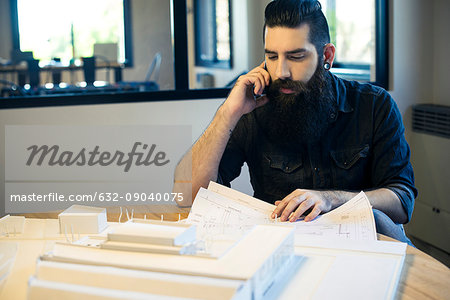 Architect looking at blueprints and talking on cell phone in office