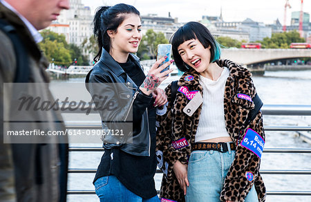 Two young stylish women laughing at smartphone on millennium footbridge, London, UK
