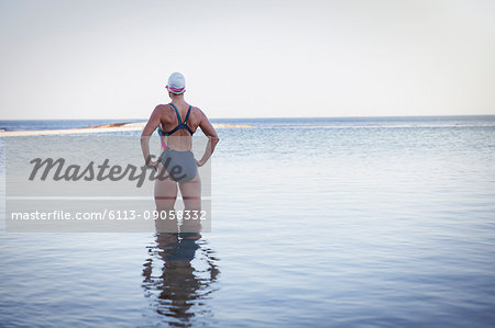 Pensive, determined female open water swimmer with hands on hips in ocean surf