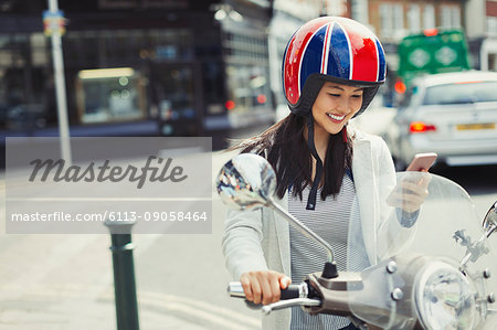 Smiling young woman texting with cell phone on motor scooter, wearing helmet on urban street