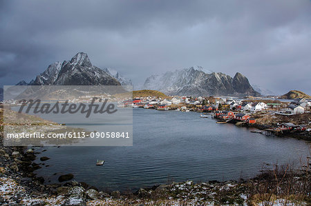 Fishing village at waterfront below snowy, rugged mountains, Reine, Lofoten, Norway