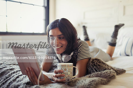 Smiling young woman drinking coffee and using digital tablet on bed