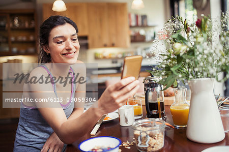 Smiling young woman texting with smart phone at breakfast table