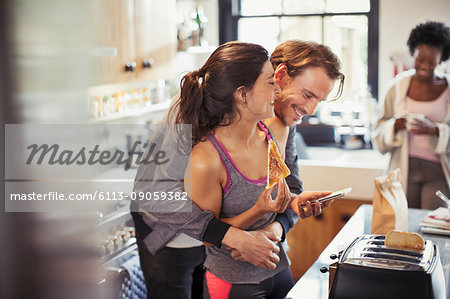 Playful couple hugging, texting with smart phone and eating toast