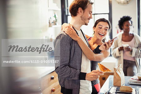 Affectionate couple hugging, texting with smart phone in kitchen
