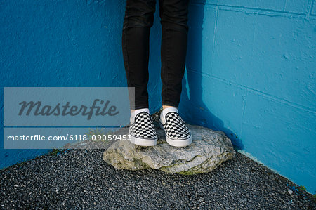 Teenage girl wearing black trousers and checkered canvas shoes, standing on rock against blue wall.