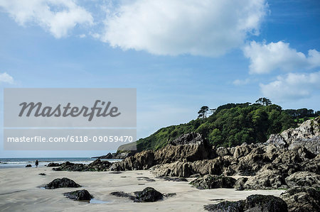 The headland and rocks on a sandy beach on the coastline under a blue sky with light cloud. A single person walking.