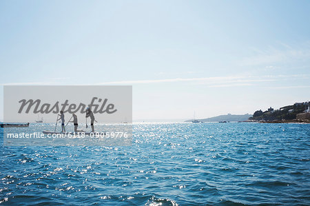 Silhouette of three people on paddleboards on the water, view out to sea from a sheltered cover to moored yachts and houses on the headland.