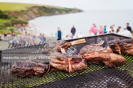 A beach barbeque with meat cooking over an open fire. People in the background on the cliffs overlooking the coastline.