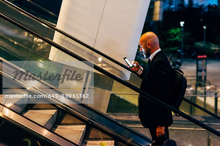 Mature businessman outdoors at night, on escalator, using smartphone