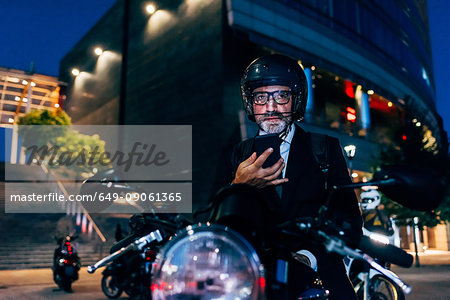 Mature businessman outdoors at night, sitting on motorcycle, using smartphone