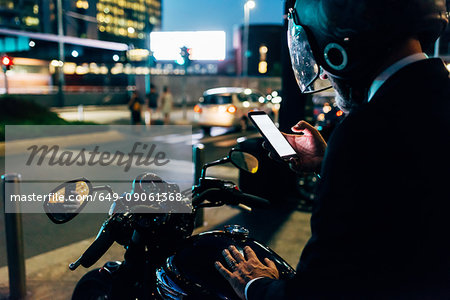 Mature businessman outdoors at night, sitting on motorcycle, wearing motorcycle helmet, using smartphone, rear view