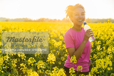 Outdoor portrait of beautiful happy mixed race African American girl teenager female young woman athlete runner drinking water from a bottle in a field of yellow flowers at sunset in golden evening sunshine