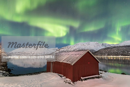 The Northern Lights (aurora borealis) reflected in the icy sea frame a typical Rorbu, Manndalen, Kafjord, Lyngen Alps, Troms, Norway, Scandinavia, Europe