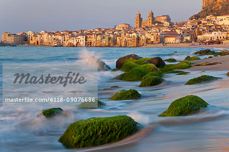 View along coastline with sandy beach, rocks covered in green algae, city in the distance.