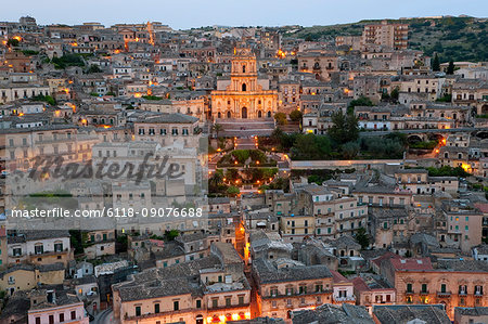 High angle view over illuminated rooftops of traditional houses in a Mediterranean city at dusk, church in the centre.