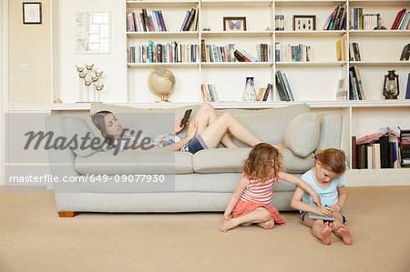 Pregnant woman reclining on sofa with smartphone and daughters sitting on floor drawing