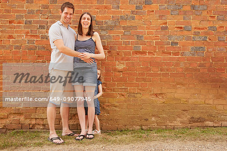 Portrait of pregnant mid adult couple and hiding daughter by brick wall