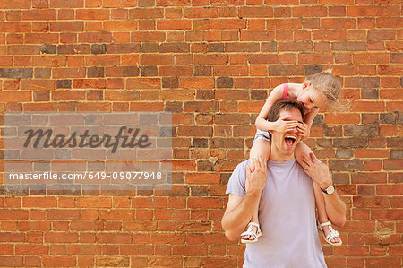 Girl getting shoulder carry from father with covering his eyes by brick wall