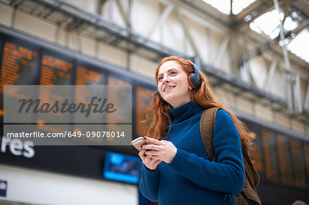 Young woman at train station, wearing headphones, holding smartphone