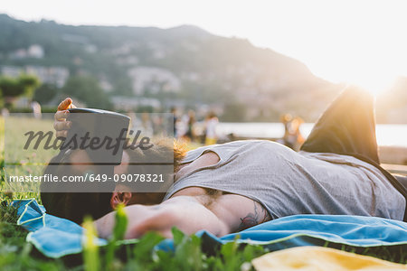 Young male hipster lying on grass looking at virtual reality headset, Lake Como, Lombardy, Italy
