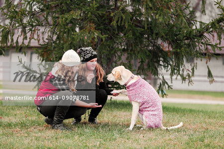 Girl and her sister crouching to hold dog's paw in garden