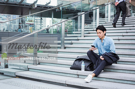 Businessman wearing blue shirt sitting outdoors on steps, looking at mobile phone.