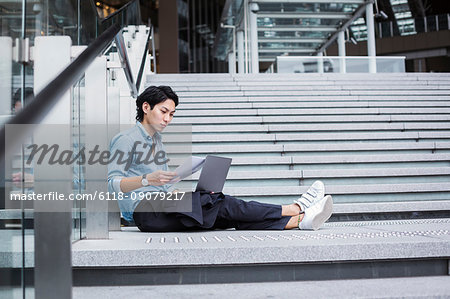 Businessman wearing blue shirt sitting outdoors on steps, holding papers, laptop on his knees.