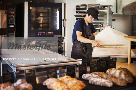 Man working in a bakery, preparing large tray with dough for rolls, oven in the background.
