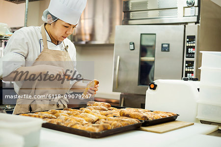 Woman wearing chef's hat and apron working in a bakery, slicing freshly baked rolls on large trays.