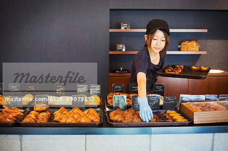 Woman working in a bakery, placing freshly baked croissants and cakes on large trays on a counter.
