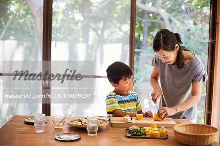 Woman and boy standing at a table, preparing corn on the cob, smiling.