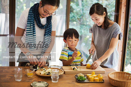 Man, woman and boy standing at a table, preparing corn on the cob, smiling.