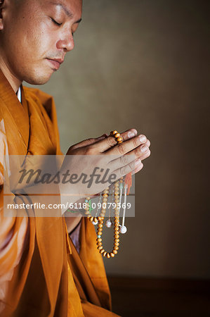 Side view of Buddhist monk with shaved head wearing golden robe kneeling indoors in a temple, holding mala, praying.