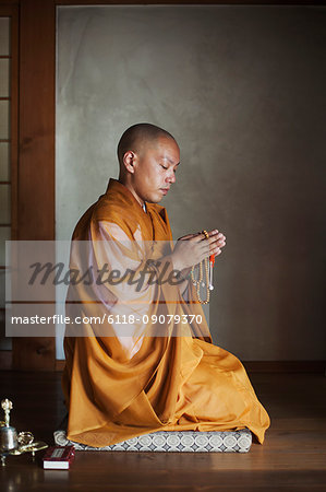 Side view of Buddhist monk with shaved head wearing golden robe kneeling indoors in a temple, holding mala, praying.