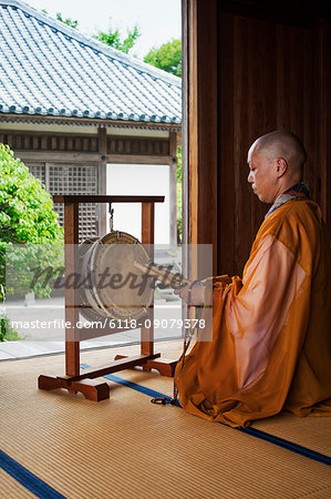 Side view of Buddhist monk with shaved head wearing golden robe kneeling in front of drum inside temple.