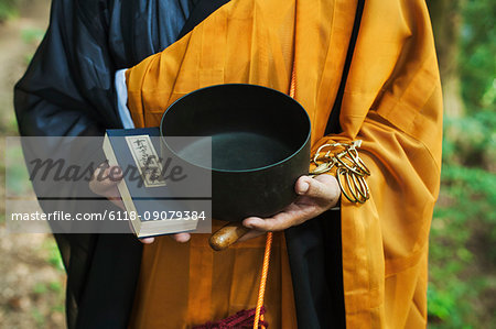 Close up of Buddhist monk wearing black and yellow robe, standing outdoors, holding prayer book and singing bowl.