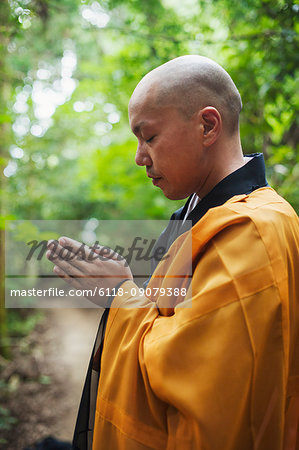 Side view of Buddhist monk with shaved head wearing black and yellow robe, standing outdoors, meditating.