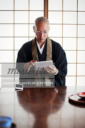 Buddhist monk with shaved head wearing black robe sitting indoors at a table, using digital tablet.