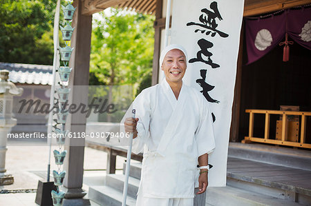 Buddhist monk wearing white robe and cap standing outside a temple, smiling at camera.