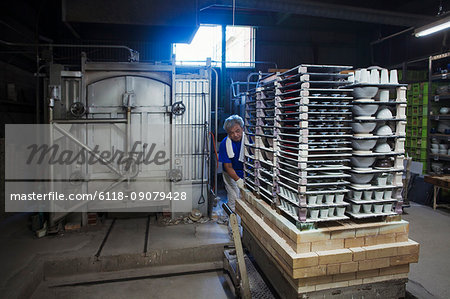 Interior view of Japanese porcelain workshop, man preparing large stack of porcelain objects for kiln.