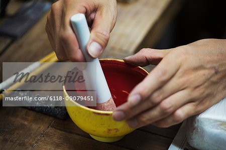 Close up of person working in a Japanese porcelain workshop, mixing red paint with pestle and mortar.