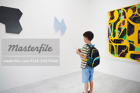 Boy with short black hair wearing backpack standing in art gallery, holding pen and paper, looking at modern painting.