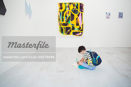 Boy with short black hair wearing backpack sitting on floor in art gallery with pen and paper, looking at modern painting.