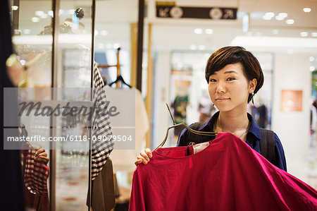 Woman with black hair wearing blue shirt standing indoors, looking at clothing in a shop, smiling at camera.