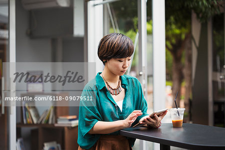 Woman with black hair wearing green shirt sitting at table in a street cafe, holding digital tablet.