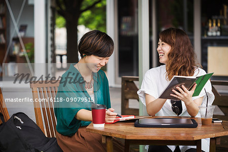 Two women with black hair wearing green and white shirt sitting at table in a street cafe, holding digital tablet, smiling.
