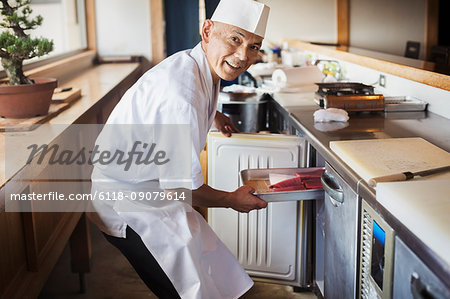 Chef working at a counter at a Japanese sushi restaurant, putting metal tray with fish in refrigerator.
