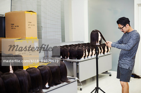 Bearded man wearing glasses standing indoors, cutting hair of brown wig on mannequin head.